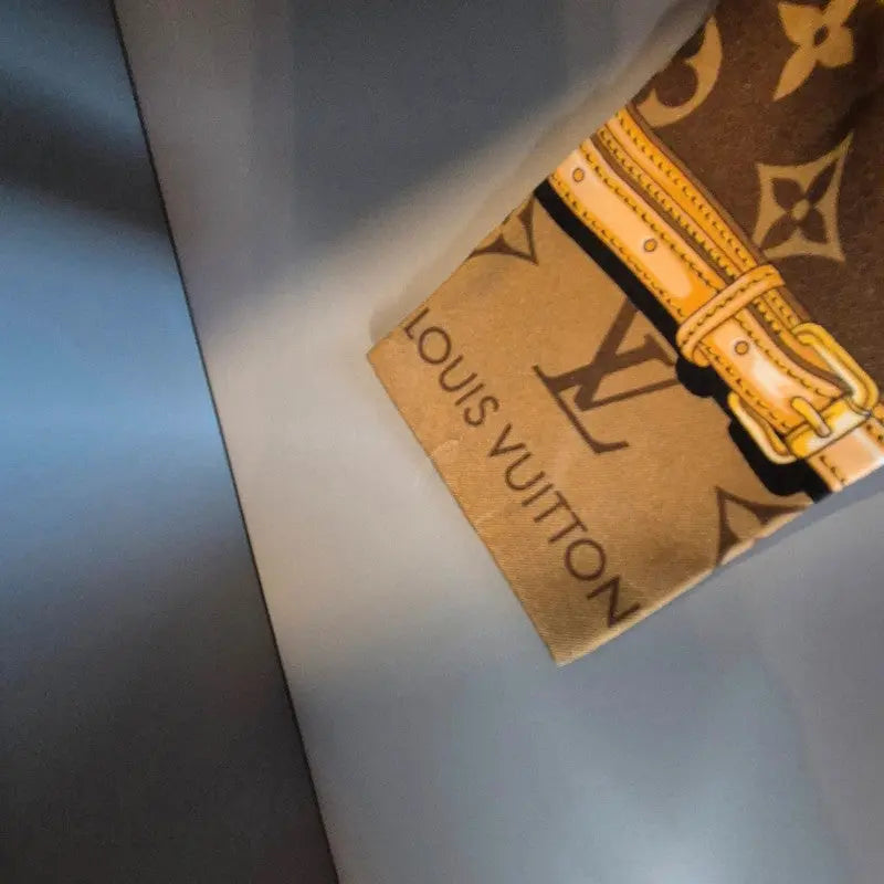 Extrémité du bandeau Confidentiel – détail boucle jaune et logo Louis Vuitton sur fond beige